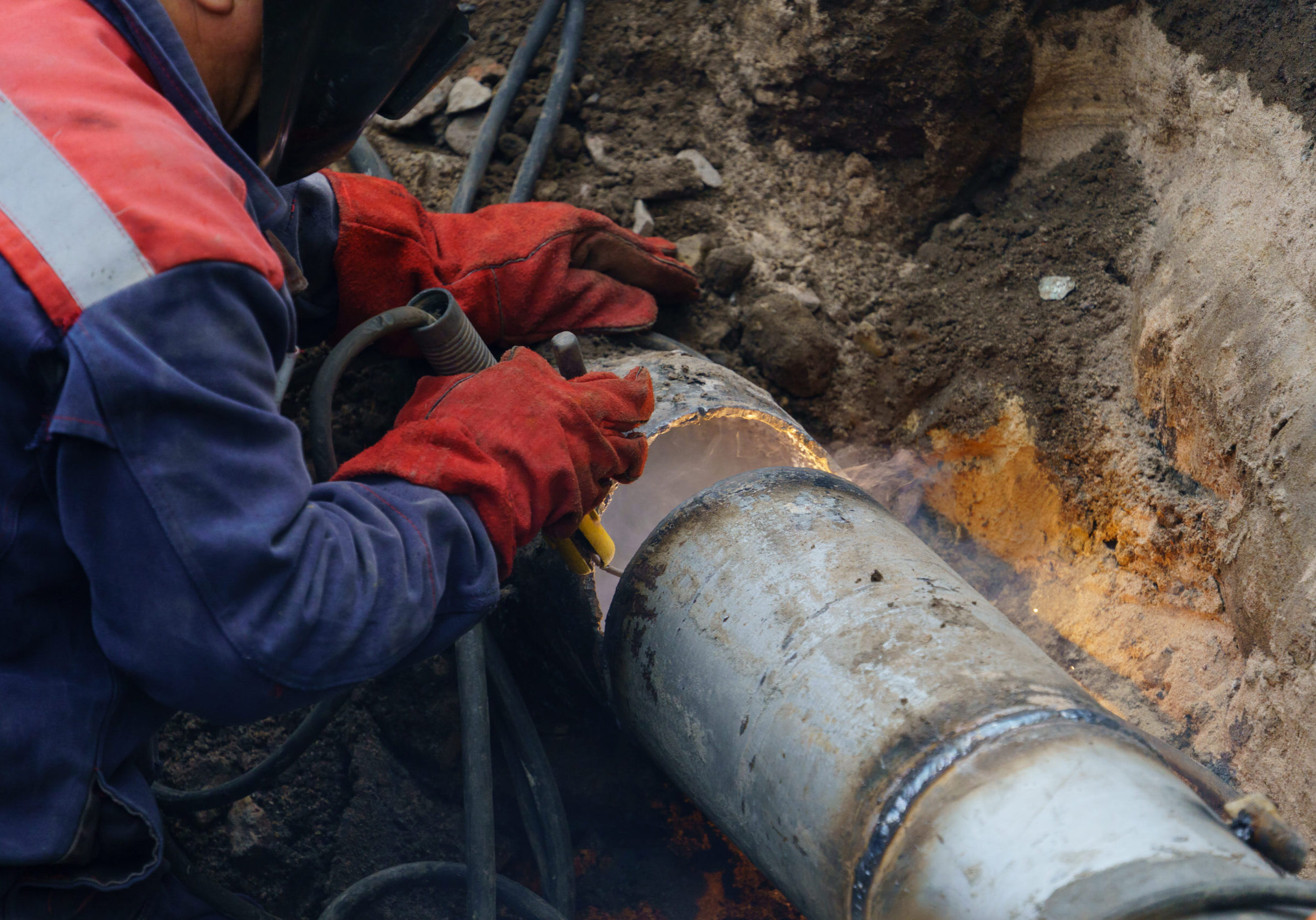 Plumber standing in pit is engaged in elimination of communal accident outdoors. Utility worker fixing broken water main. Welding work to replace rotten elements of water pipe. Sewerage pipe repair